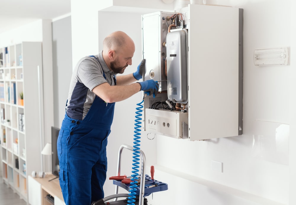 An HVAC technician services an open wall-mounted boiler using an air hose.